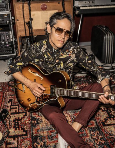 Damon Castillo sitting on a rug in the studio playing a guitar.